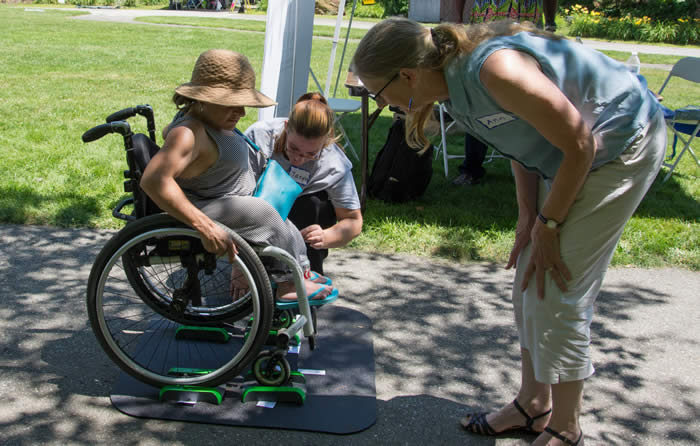 Woman in wheelchair on portable scale mat, outdoors, another woman is helping. 
